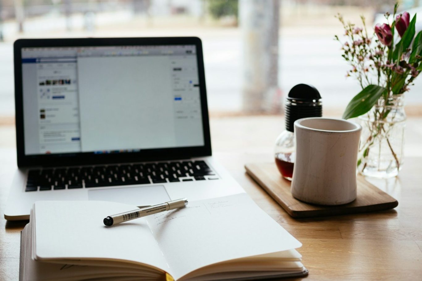 Webinare Laptop mit geöffnetem Notizbuch, Kaffeetasse und Blumen auf einem Tisch.