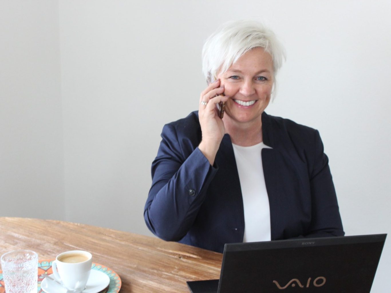 Videocall Lächelnde Frau mit Handy am Tisch, Laptop und Kaffeetasse sichtbar.